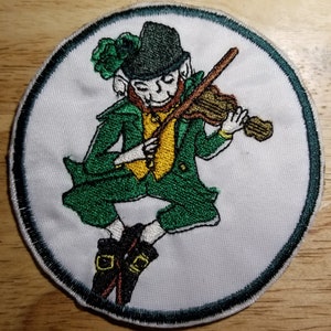 Op de afbeelding: Gestikte patch met een groene kabouter die viool speelt. De kabouter draagt een groene hoed, een groen vest en een groene broek. De patch is rond en heeft een groene rand.