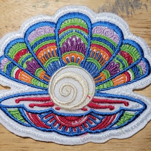 May include: Embroidered seashell patch with a pearl. The patch features a white border and a colorful design with blue, green, orange, purple, and red details. The pearl is a cream color with a spiral design.