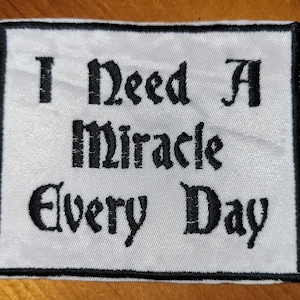 May include: White embroidered patch with black text that reads "I Need A Miracle Every Day".