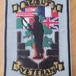 May include: A gray embroidered patch with a silhouette of a soldier standing in front of a tree. The soldier is holding a rifle and there are flags of the United States, France, Ireland, and the United Kingdom behind the soldier. The text "BELAUT" is above the soldier and the text "VETERAN" is below the soldier.