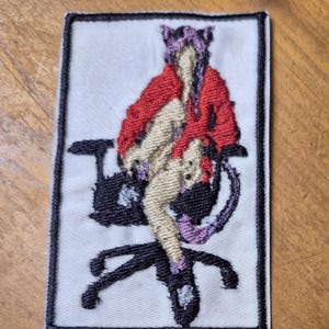 May include: Embroidered patch featuring a figure in a red jacket and purple hair, seated on a black chair. The patch has a white background with a black border. The figure's skin is beige, and the chair has purple accents.