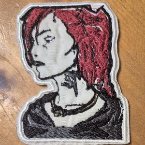 May include: Embroidered patch featuring a stylized portrait of a person with red hair and a black outfit. The patch has a white border and is set against a wooden surface. The design includes a necklace and facial details.