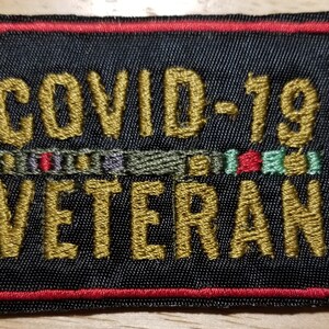 May include: A rectangular black patch with gold embroidered text reading "COVID-19 VETERAN." The patch has a red border and a row of colorful embroidered ribbons. A military-themed item.