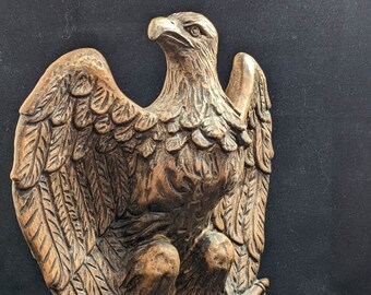 May include: A bronze eagle sculpture with its wings spread wide, perched on a draped fabric base. The eagle's head is turned to the side, and its talons are gripping the fabric.