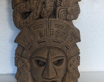 May include: A brown clay sculpture of a Mayan mask with intricate details. The mask features a serpent head on top and a human face with a large nose and wide eyes.