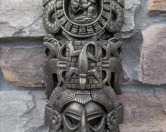 May include: A bronze-colored, three-tiered wall sculpture with Mayan-inspired designs. The top tier features a circular design with a central figure, the middle tier has a lizard and serpent motifs, and the bottom tier depicts a mask.