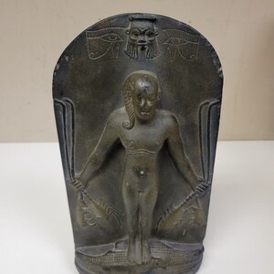 May include: A dark gray stone sculpture of a standing figure with outstretched arms. The figure is carved in a relief style and has a stylized face. The sculpture is decorated with hieroglyphs and the Eye of Horus symbol.