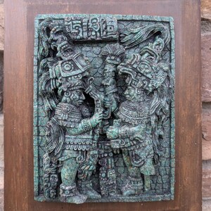 May include: A teal-colored relief carving of two figures in ornate headdresses and clothing, set within a wooden frame. The figures appear to be holding a staff or similar object. The carving has a detailed, textured surface.