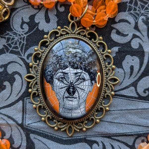 May include: An oval pendant with a vintage-style illustration of a face with a spider web design and a spider. The pendant is in a bronze-colored frame and is part of a necklace with orange beads. The background is a dark gray fabric with a spiderweb pattern.