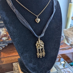 May include: Two necklaces displayed on a black velvet bust. The top necklace is a delicate gold chain with a heart pendant. The bottom necklace features a black ribbon and a gold filigree pendant with dangling elements. The jewelry is displayed on a black velvet bust.