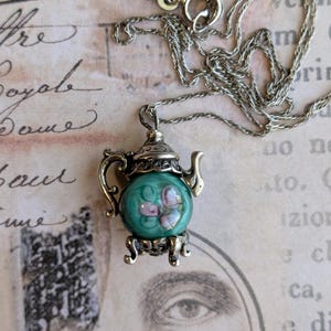 May include: A silver-toned metal necklace featuring a teapot-shaped pendant. The pendant has a round, translucent green glass center with a painted pink floral design, a decorative lid, a curved handle, a spout, and four small ornate feet.