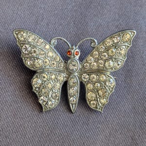 May include: A silver-toned butterfly brooch encrusted with clear rhinestones. The butterfly has orange-red eyes and is set against a purple background. The brooch is a classic jewelry piece.