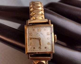 Può includere: Un orologio da polso dorato con un quadrante quadrato e un cinturino in metallo dorato. Il quadrante dell'orologio ha indici orari in numeri romani neri e il testo "Lady Elgin" stampato su di esso.