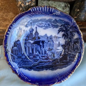 May include: A blue and white porcelain bowl with a gold rim. The bowl features a detailed scene of a castle and palm trees.