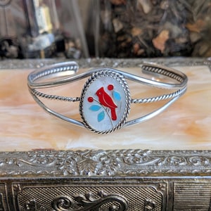 May include: A silver bracelet with a white oval charm featuring a red cardinal bird with turquoise accents. The bracelet has a twisted wire design.