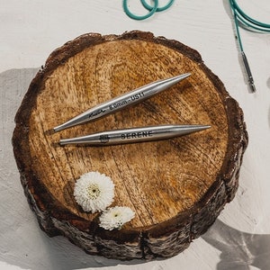 Könnte beinhalten: Zwei silberne Stricknadeln auf einer Holzoberfläche. Die Nadeln sind mit "Knitter 8,0 mm - US11" und "SERENE" beschriftet.