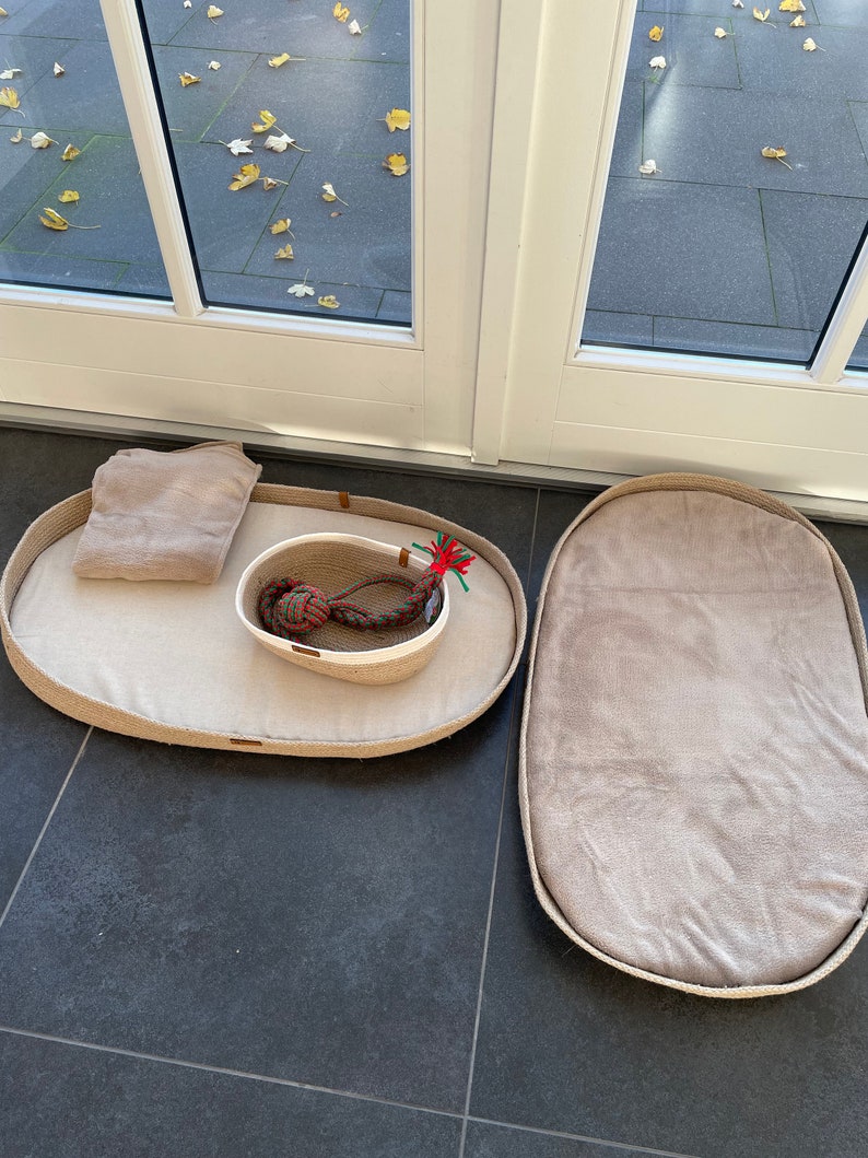 May include: Two oval-shaped pet beds made of natural jute rope with a beige fabric lining. One bed has a small white bowl with a red and green rope toy inside.