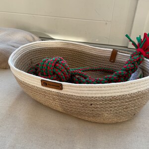 May include: A woven jute basket with a white stripe and a brown leather tag that says "Handmade". The basket contains a red and green rope toy.