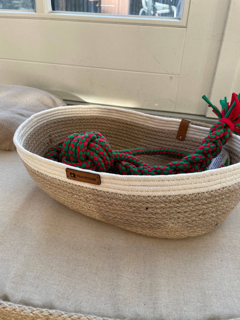 May include: A woven jute basket with a white stripe and a brown leather tag that says "Handmade". The basket contains a red and green rope toy.