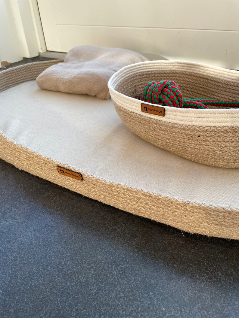 May include: A beige oval-shaped pet bed with a brown "Handmade" label. The bed is made of woven rope and has a soft, white fabric lining. A smaller, round woven rope basket with a white rim and a green and red rope toy inside is sitting on top of the bed. The basket also has a brown "Handmade" label.