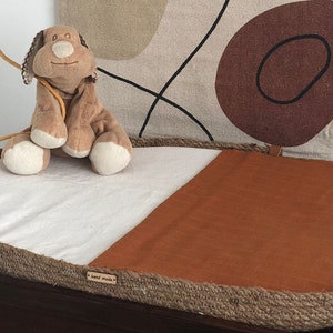 May include: An oval-shaped changing mat with a rope border. The mat is divided in half, with one side white and the other side brown. A small stuffed dog toy is sitting on the white side of the mat. The mat has a "hand made" label on the bottom.