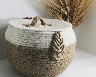 May include: A round, woven storage basket with a lid. The basket is made of natural fibers and has a light brown and white color scheme. The lid has a rope handle.