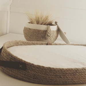 Puede incluir: Una cuna redonda tejida de fibra natural con un forro de tela blanco. La cuna tiene una correa de cuero marrón con una X. Una cesta redonda más pequeña, tejida con un borde blanco, se coloca encima de la cuna.