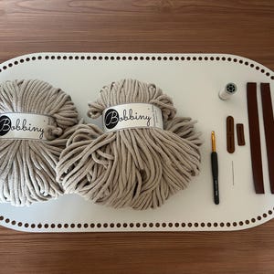 May include: A craft kit with two beige yarn balls labeled "Bobbini" on a white oval tray. Includes a crochet hook, needle, thread, and leather straps. Ideal for crochet projects.