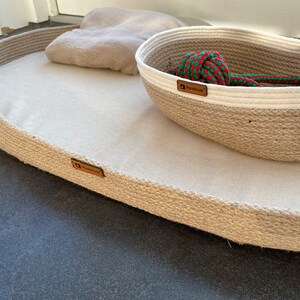 May include: A beige oval-shaped pet bed with a brown "Handmade" label. The bed is made of woven rope and has a soft, white fabric lining. A smaller, round woven rope basket with a white rim and a green and red rope toy inside is sitting on top of the bed. The basket also has a brown "Handmade" label.