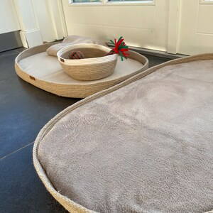 May include: Two oval-shaped pet beds made of natural rope with a soft, beige fabric lining. The smaller bed has a woven rope basket with a white rim inside.