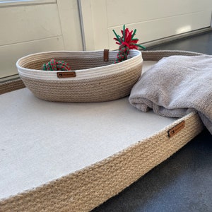 May include: A rectangular dog bed made of natural jute rope with a white fabric insert. The bed has a brown leather tag that says "Handmade". A smaller, oval-shaped jute rope basket with a white fabric rim sits on the bed. The basket contains a red and green rope toy.