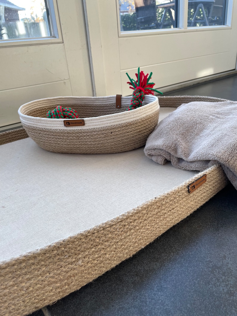 May include: A rectangular dog bed made of natural jute rope with a white fabric insert. The bed has a brown leather tag that says "Handmade". A smaller, oval-shaped jute rope basket with a white fabric rim sits on the bed. The basket contains a red and green rope toy.