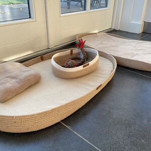May include: Two oval-shaped pet beds made of natural rope with a beige fabric lining. The smaller bed has a white and beige rope basket inside with a red, green, and white rope toy.