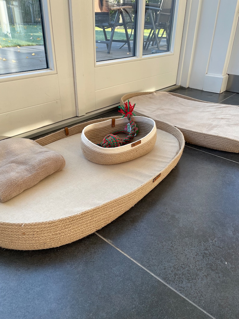 May include: Two oval-shaped pet beds made of natural rope with a beige fabric lining. The smaller bed has a white and beige rope basket inside with a red, green, and white rope toy.
