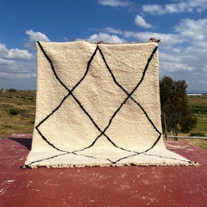 Op de afbeelding: Crèmekleurig, handgeweven vloerkleed met een vet zwart geometrisch ruitpatroon. Het vloerkleed heeft een dikke, getextureerde pool en een gefranjerde rand. Het vloerkleed wordt buiten gepresenteerd tegen een achtergrond van een blauwe lucht en een groen landschap.