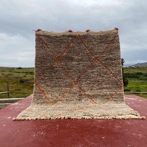 May include: A rectangular, hand-woven rug with a neutral color palette. The rug features a textured, shaggy pile in shades of beige and brown, with a geometric diamond pattern in orange. The rug is displayed outdoors on a red surface.