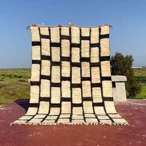 May include: A large, rectangular rug with a geometric pattern of black rectangles on a cream-colored background. The rug is displayed outdoors, held up against a blue sky and green landscape. The rug's edges have a fringe.