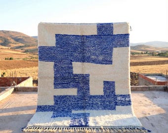 Puede incluir: Una alfombra rectangular grande con un fondo crema y un llamativo patrón geométrico azul. El diseño presenta formas y líneas interconectadas. La alfombra tiene flecos a lo largo del borde inferior y se exhibe al aire libre.