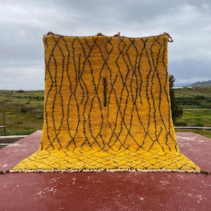 May include: A large, rectangular rug with a vibrant yellow background and a black geometric pattern. The rug has a textured appearance and is displayed outdoors, showcasing its size and design. The edges are finished with a decorative trim.