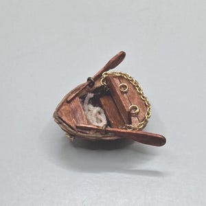 May include: A miniature wooden rowboat brooch featuring a brown hull, two attached oars, and a gold-toned metal chain draped across the interior bench. A small coil of white rope sits inside the boat, which is finished with metal hardware.