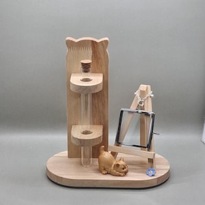 May include: A wooden display featuring a test tube vase with a cork stopper, a small wooden easel with a silver necklace, and a carved wooden cat figurine. The base is oval-shaped, and a small blue flower is placed near the easel.