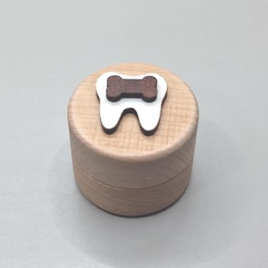 Puede incluir: Pequeña caja redonda de madera clara con tapa. La tapa está decorada con un diente blanco y un hueso marrón. La caja está hecha de madera clara.