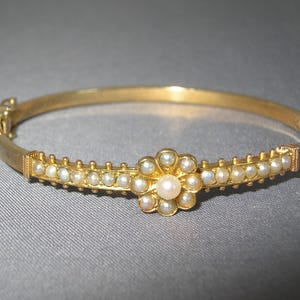 May include: A gold-toned hinged bracelet with a floral centerpiece. The bracelet features a row of small pearls and a central flower design with a larger pearl in the middle. The bracelet is approximately 7 inches in circumference.