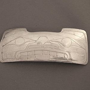 May include: A silver-colored, rectangular belt buckle with a detailed, engraved design of a stylized face. The buckle features a symmetrical pattern with circular and rectangular elements, likely representing a Native American or indigenous art style.