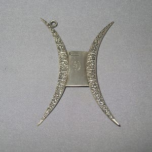 May include: A silver-toned, ornate picture frame with a unique, curved design. The frame features intricate floral patterns and a hinged back. The frame is designed to hold a small photograph or artwork, adding a touch of elegance to any display.