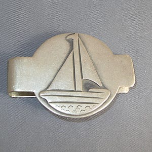 May include: Silver-toned metal money clip featuring a sailboat design. The clip has a rectangular shape with a curved top section that displays the sailboat. The sailboat has a triangular sail and a detailed hull.