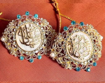 May include: Two circular gold-tone brooches featuring intricate filigree metalwork, clear rhinestones, and small turquoise-colored stones. Each brooch has a central white disc with gold-tone Arabic calligraphy and is attached to a gold cord.