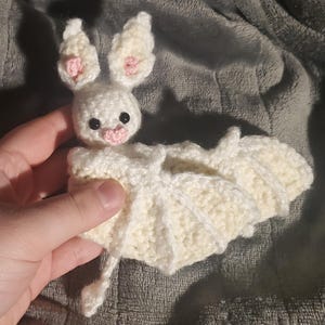 Puede incluir: Un murciélago blanco de crochet con orejas rosas y una nariz rosa. El murciélago está acostado sobre una superficie gris.