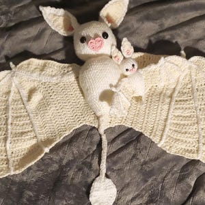 Puede incluir: Un murciélago blanco de crochet con una nariz rosa y dos murciélagos más pequeños colgando de sus alas. Los murciélagos están sobre una superficie gris texturizada.