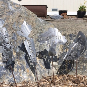 May include: Five metal garden markers with different vegetable names. The markers are shaped like the vegetables they represent. The names are "Brussels Sprouts", "Carrot", "Broccoli", "Melon", and "Watermelon".